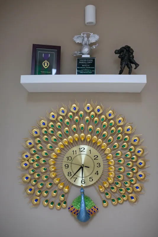 Decorative peacock wall clock below a shelf displaying a Purple Heart and a United States Marine Corps plaque