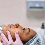 a woman in profile lies down with a hair covering while someone with gloves holds her face.