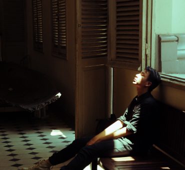 a young man sits on a bench in a dark room, with light slanting across his face.